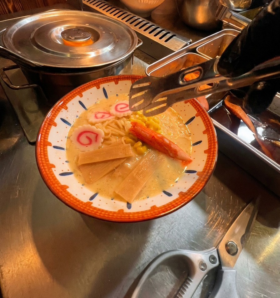 Pinzas colocando ingredientes coloridos como pasteles de pescado rosados, un palito de cangrejo y maíz en un tazón de ramen.