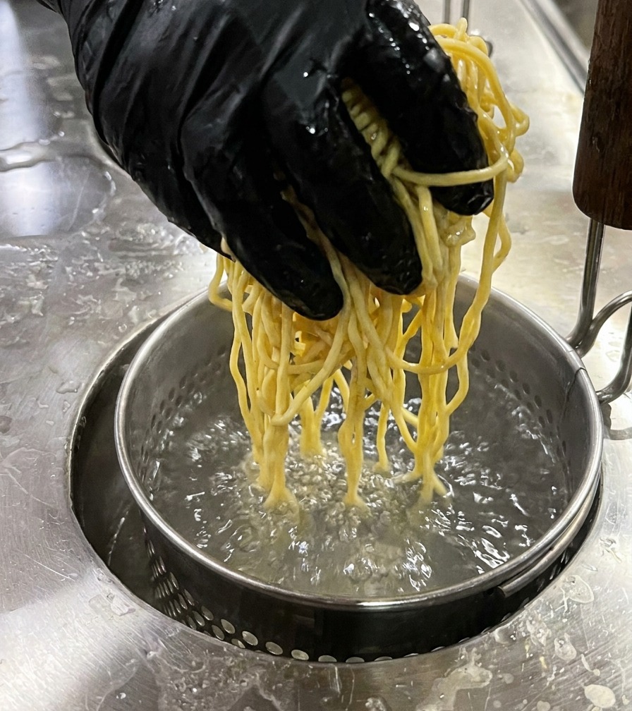 Una mano bajando un colador metálico con fideos amarillos en agua hirviendo.