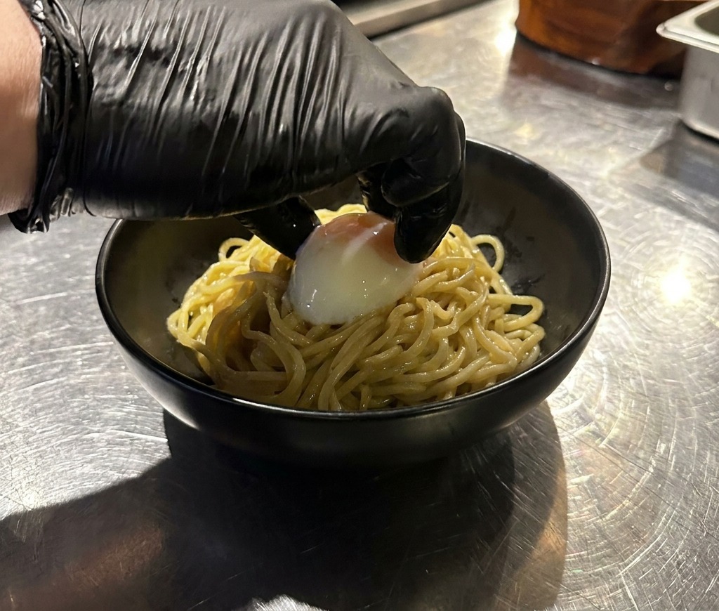 Una mano colocando un huevo onsen sobre una montaña de fideos ramen en un bol negro.
