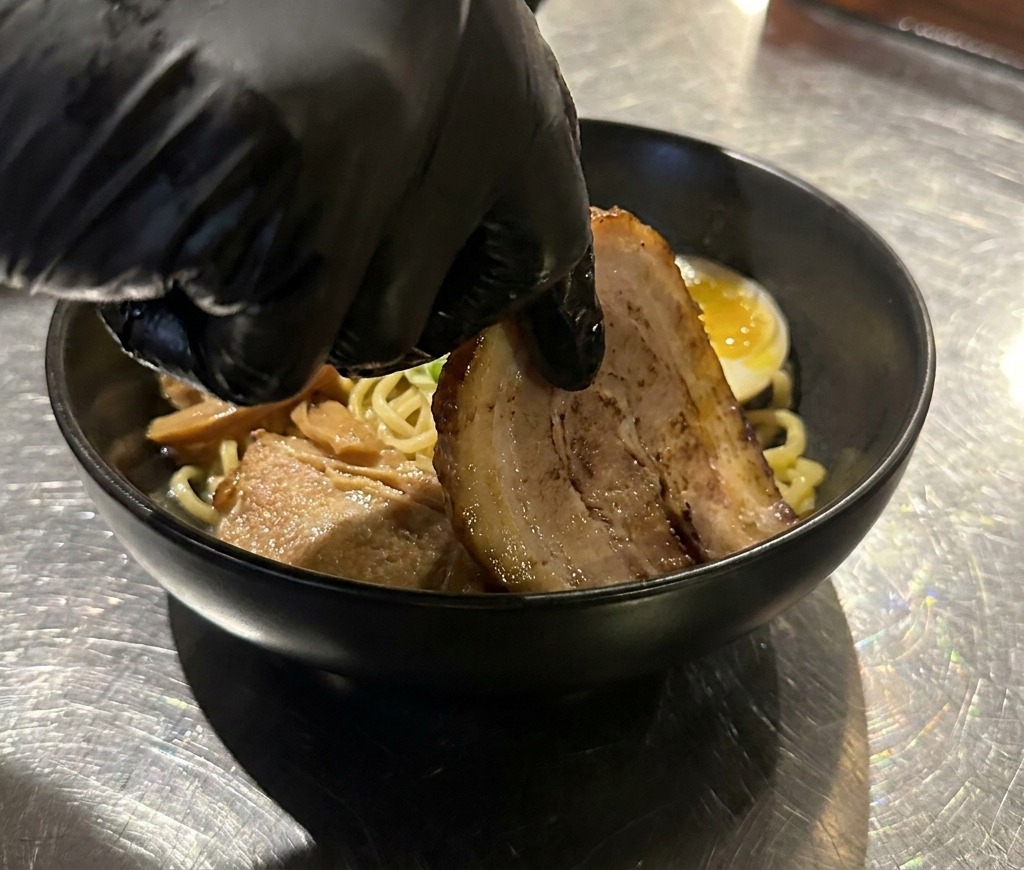 Una mano colocando una rodaja de cerdo chashu sellado en un bol de fideos ya decorado.