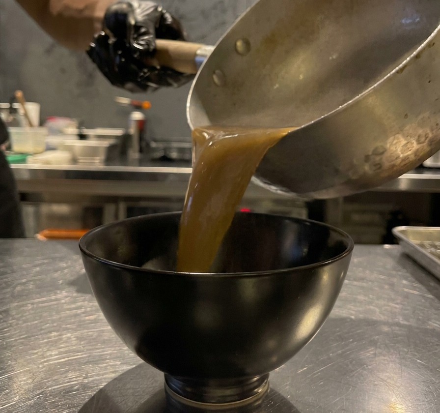 Salsa espesa de ajo negro vertiéndose desde una olla en un pequeño cuenco de cerámica negra.