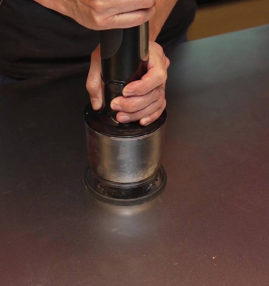 Una persona usando una batidora de mano negra para triturar ajo carbonizado dentro de un vaso metálico.