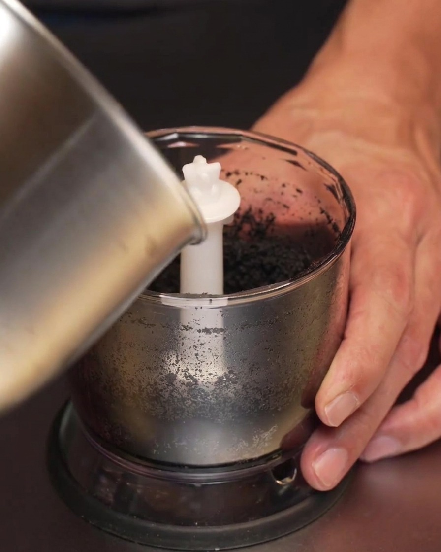 Aceite infusionado con ajo oscurecido vertiéndose desde un cazo metálico en un vaso de batidora con pasta de ajo negro.