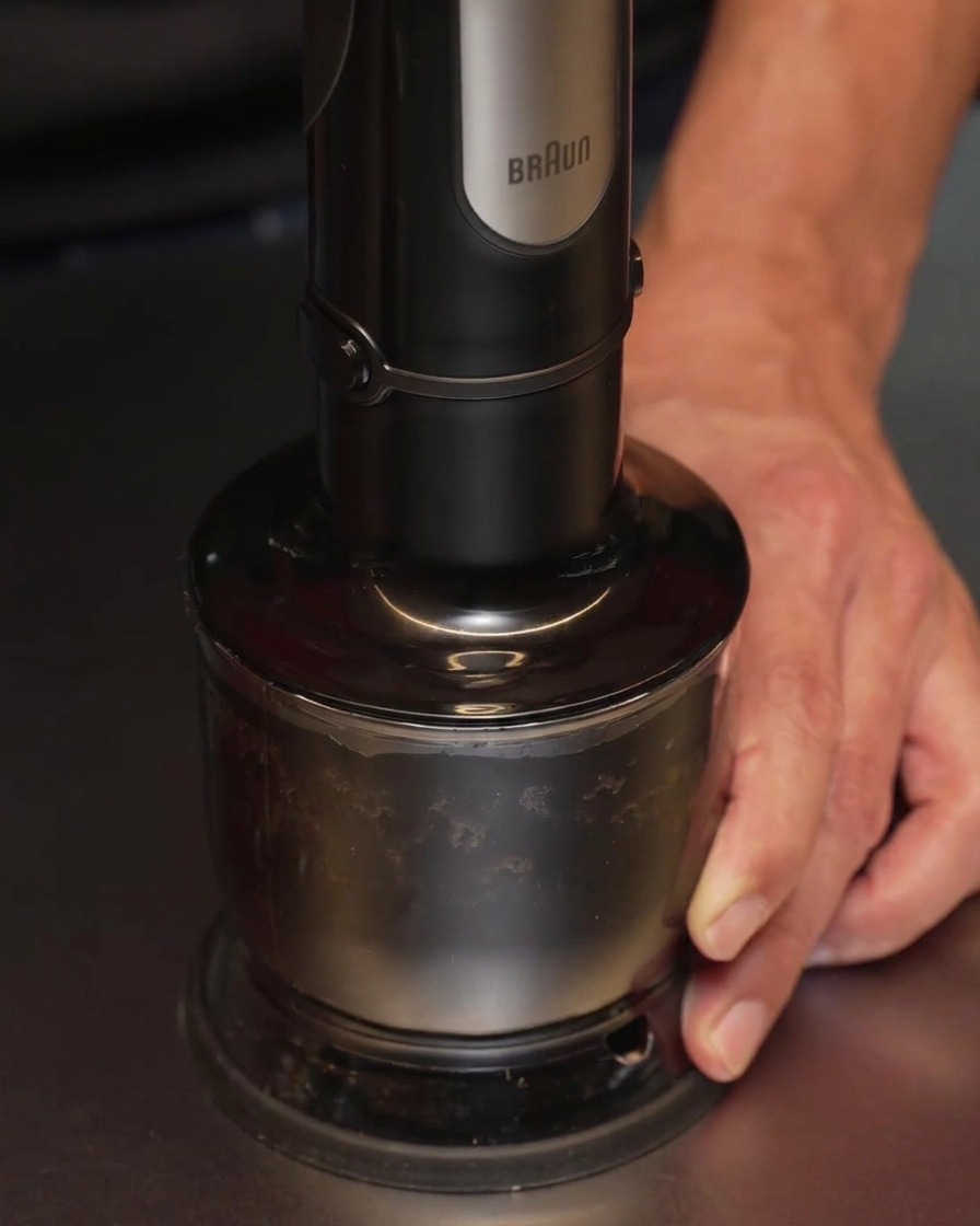 Una batidora de mano procesando ajo carbonizado y aceite en un recipiente, convirtiendo la mezcla en un líquido negro opaco.