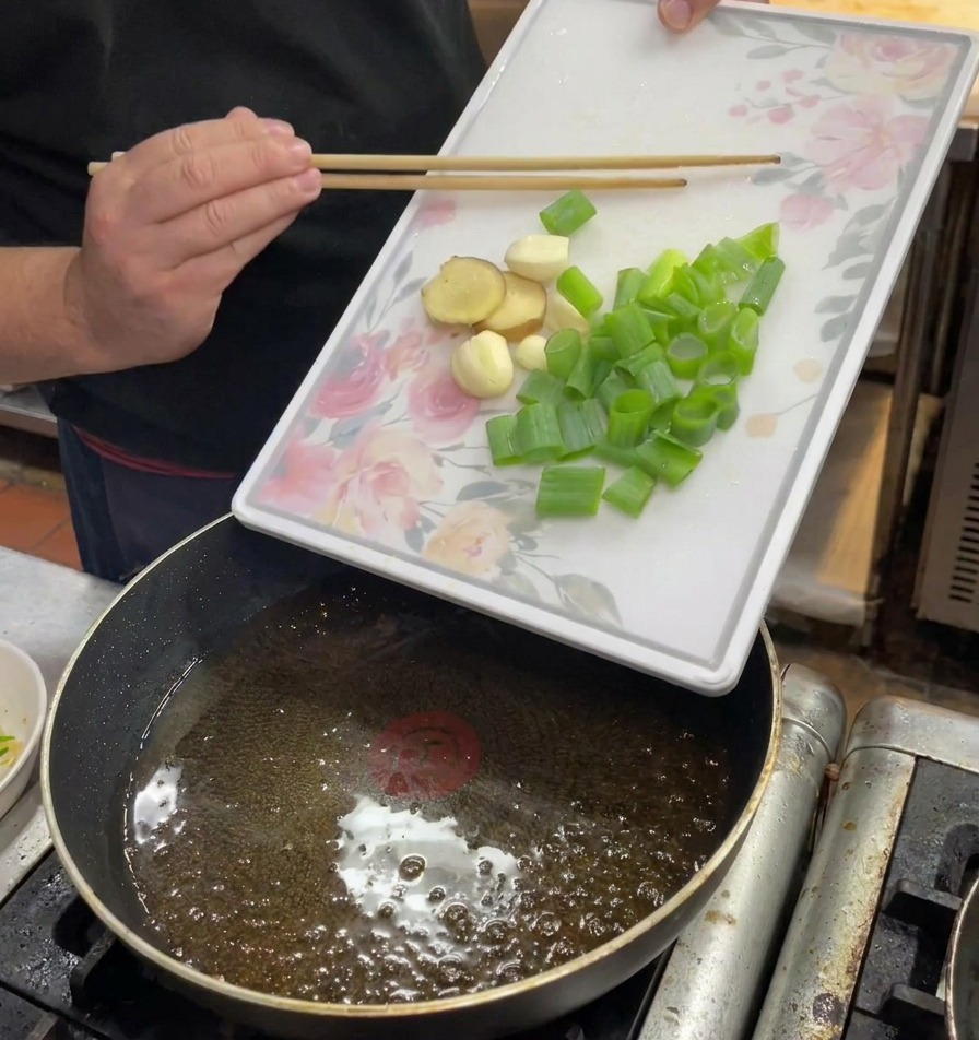 Manos deslizando cebolleta, jengibre y ajo picados desde una tabla de cortar a una sartén con aceite caliente.