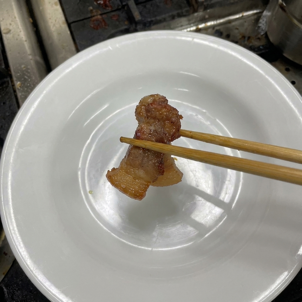 Palillos de madera sosteniendo un trozo de panceta tierna y marrón oscuro sobre un plato blanco.