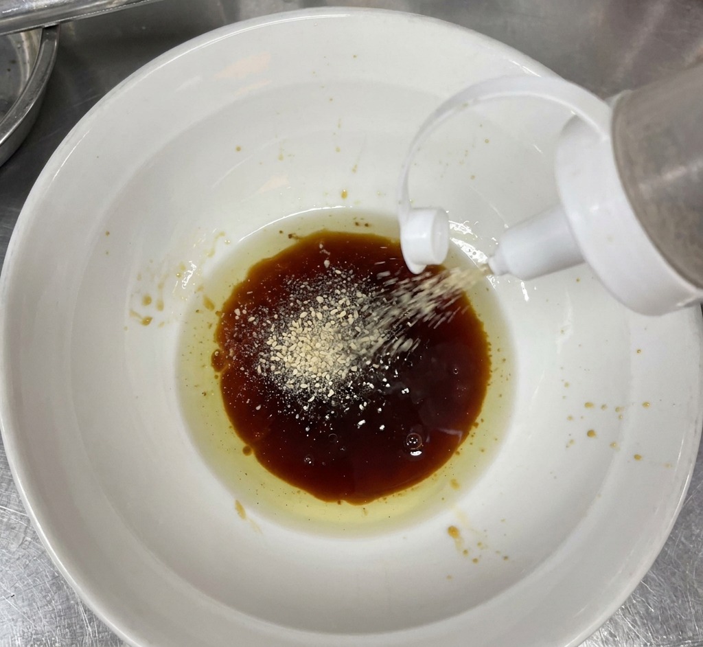 Sésamo blanco en polvo cayendo desde un biberón de cocina a un bol con salsa oscura y aceite dorado.