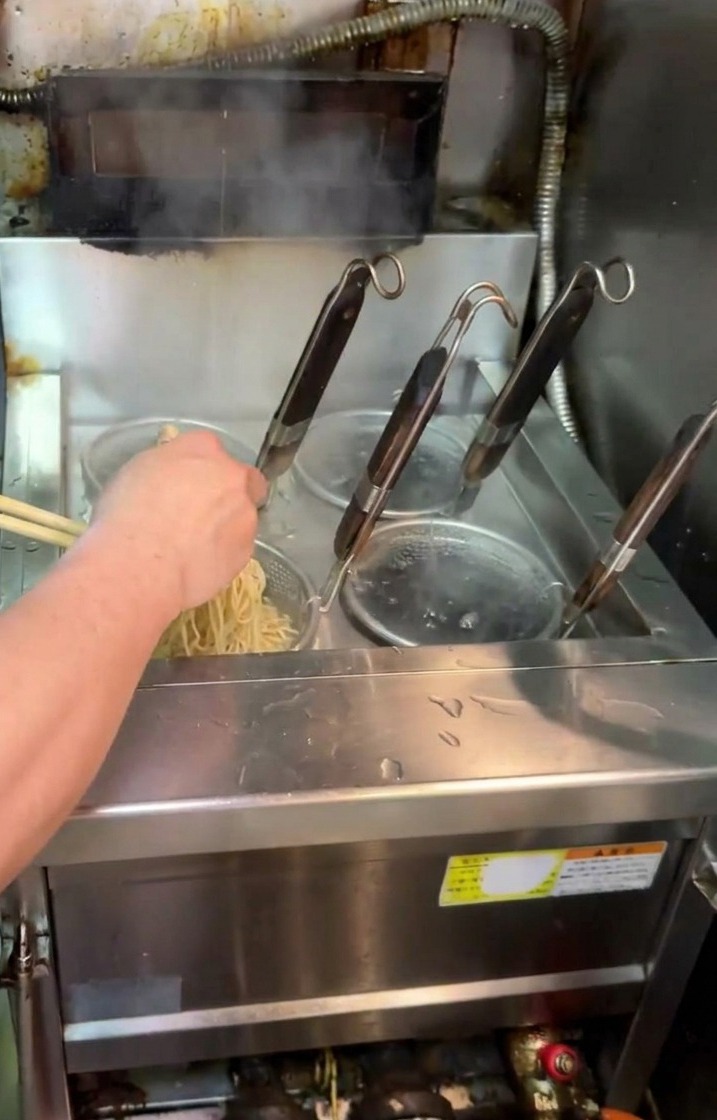 Un cocinero levantando un colador de malla con fideos ramen de una olla y sacudiéndolo para escurrir el agua.