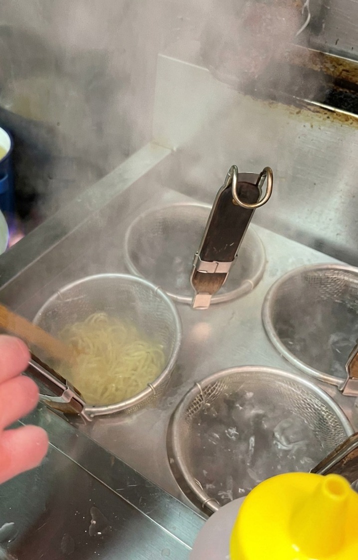 Fideos cocinándose dentro de un colador de malla sumergido en una olla con agua hirviendo en una cocina profesional.