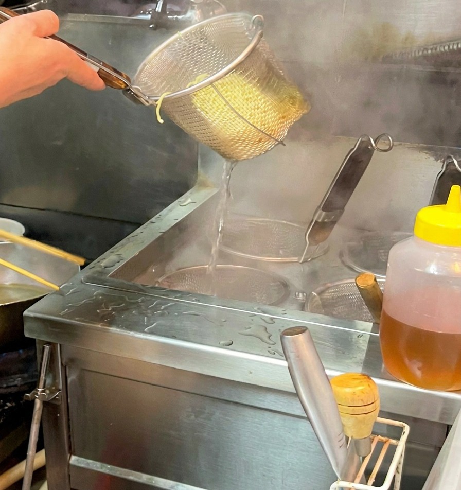 Una mano sosteniendo un colador de acero sobre un bol, soltando los fideos ramen cocidos en un caldo oscuro y humeante.