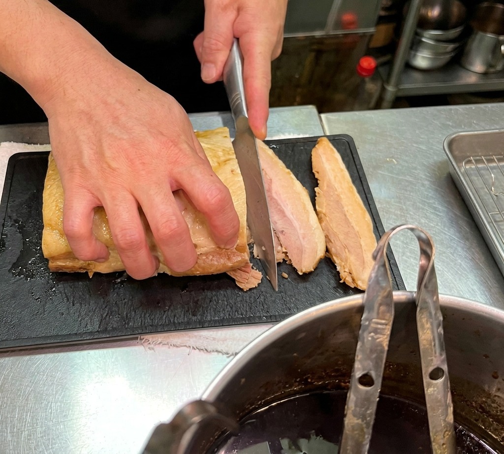 Primer plano de un chef cortando un trozo de panceta de cerdo tierna en lonchas gruesas sobre una tabla de cortar negra.