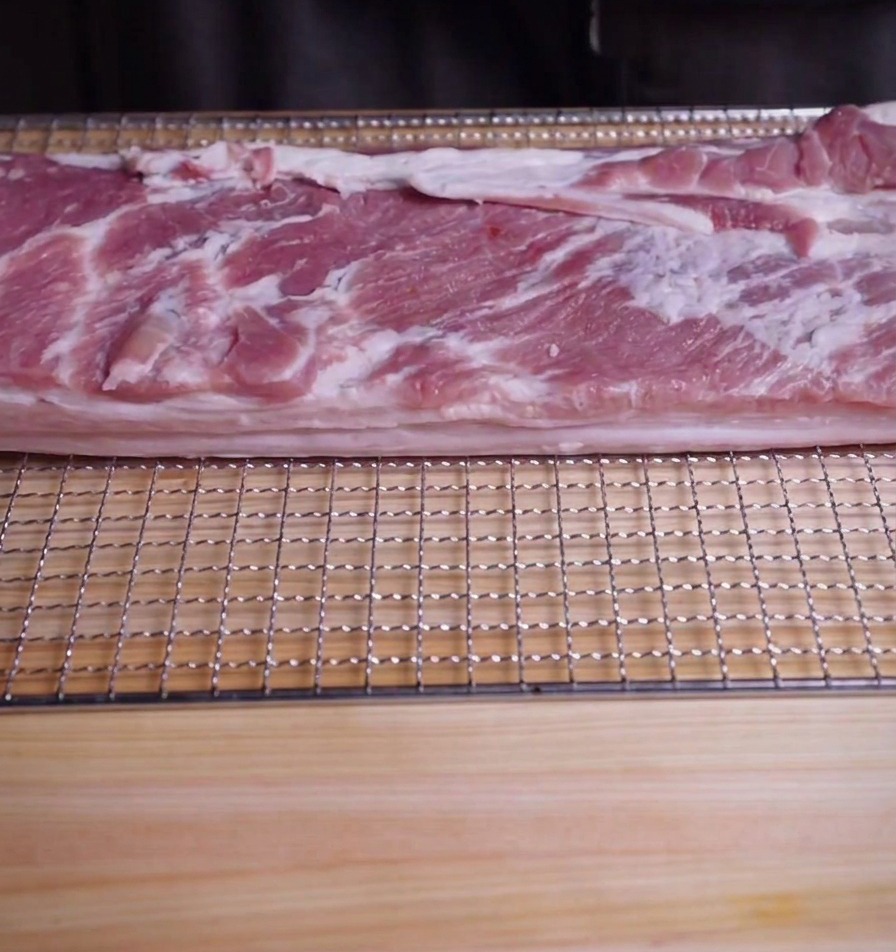 Una pieza rectangular de panceta de cerdo cruda sobre una rejilla y una tabla de madera.