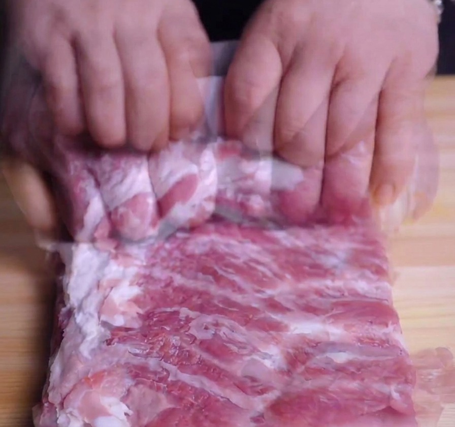 Manos enrollando con fuerza una pieza de panceta de cerdo para formar un cilindro sobre una tabla de madera.