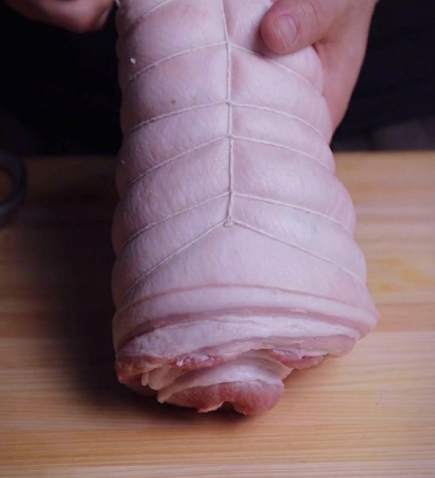 Manos sujetando un rollo de panceta mientras lo envuelven con hilo bramante blanco de cocina.
