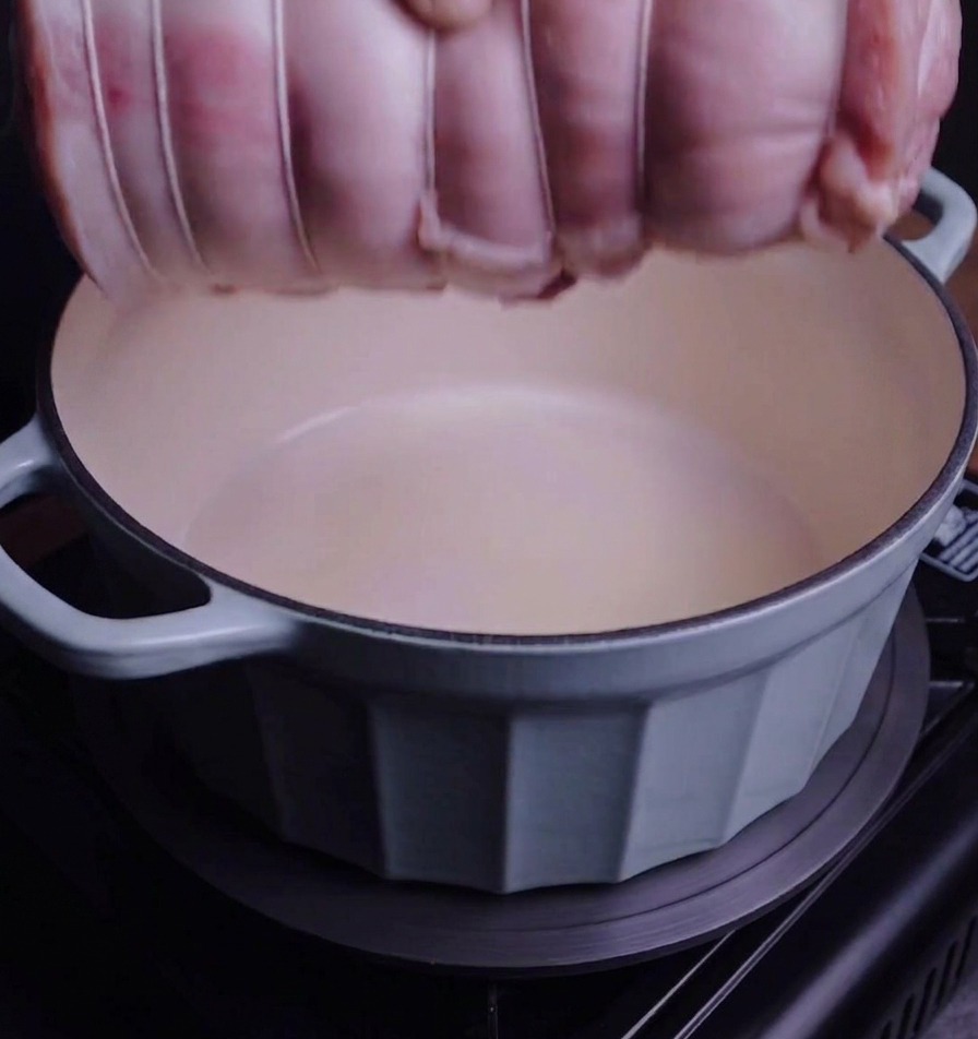 Un rollo de panceta atado siendo colocado en una olla de hierro fundido de color azul claro.