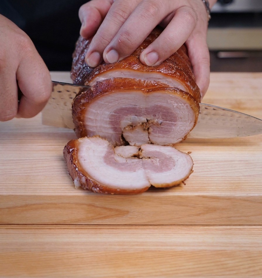 Un chef usando un cuchillo afilado para cortar rodajas redondas de un rollo de chashu frío.