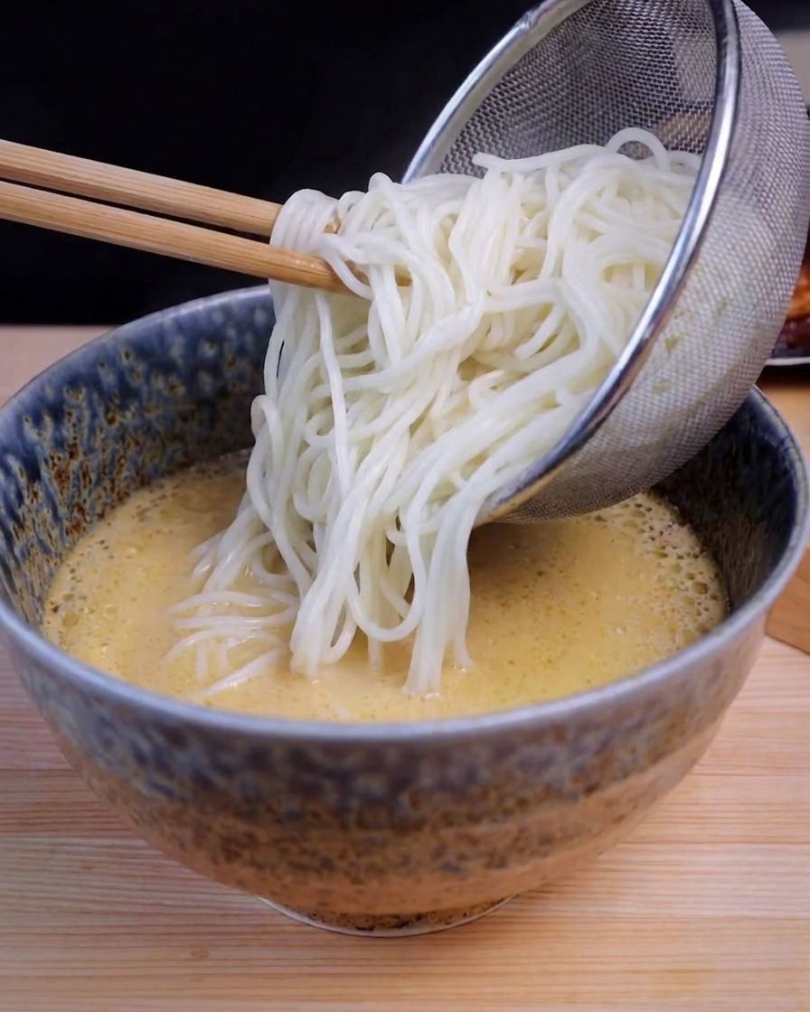 Un colador de acero inoxidable vertiendo fideos blancos en un cuenco con caldo de ramen amarillo.