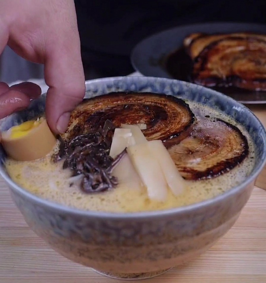 Una mano colocando medio huevo marinado en un cuenco de ramen con chashu, setas y bambú.