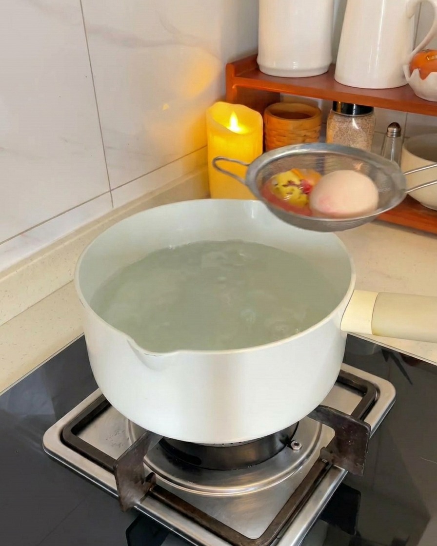 Dos huevos blancos en un colador de malla metálica sumergiéndose en una olla de agua hirviendo sobre una estufa.