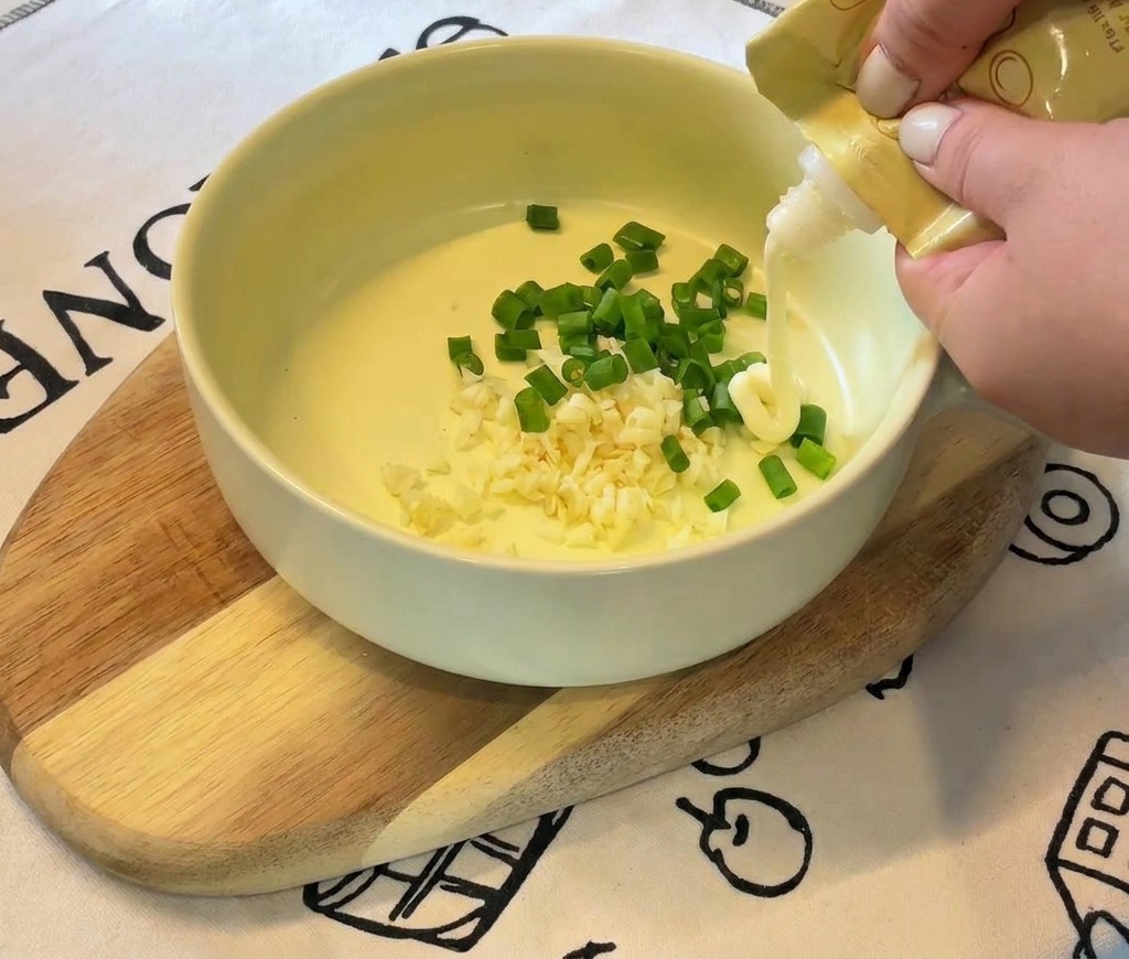 Manos exprimiendo mayonesa blanca de un envase amarillo en un tazón que contiene ajo y cebollín.