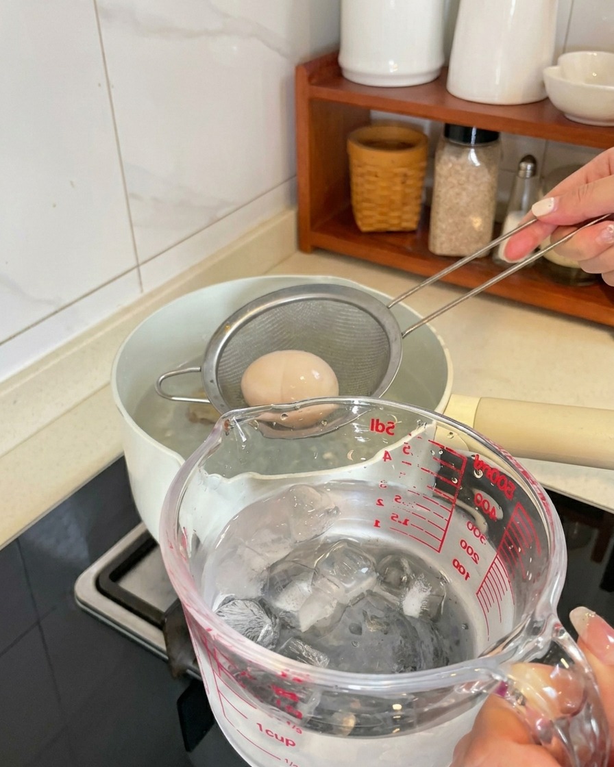 Un huevo cocido siendo levantado de la olla con un colador y colocado en una jarra con cubitos de hielo y agua.