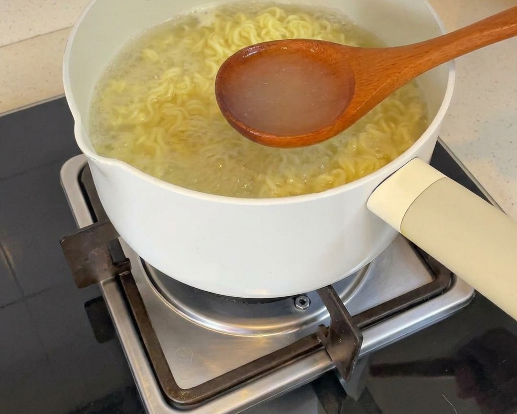 Un cucharón de madera sacando agua caliente y humeante de la cocción de los fideos en una olla blanca.