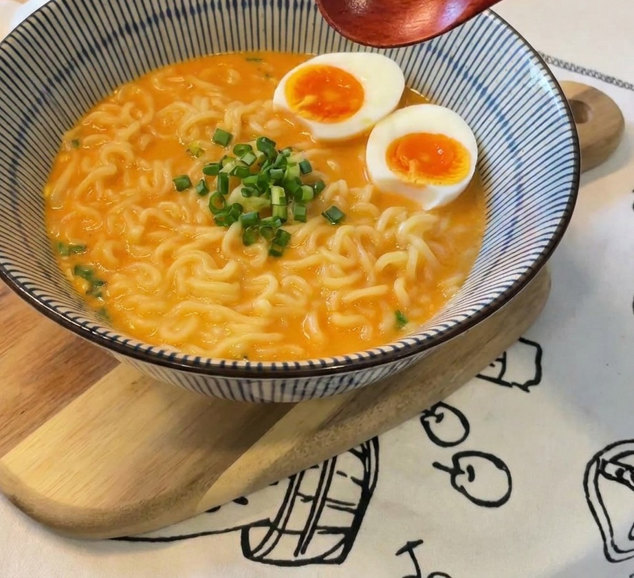 Tazón de ramen con huevos, una montaña de cebollín fresco en el centro y una cuchara de madera sostenida arriba.