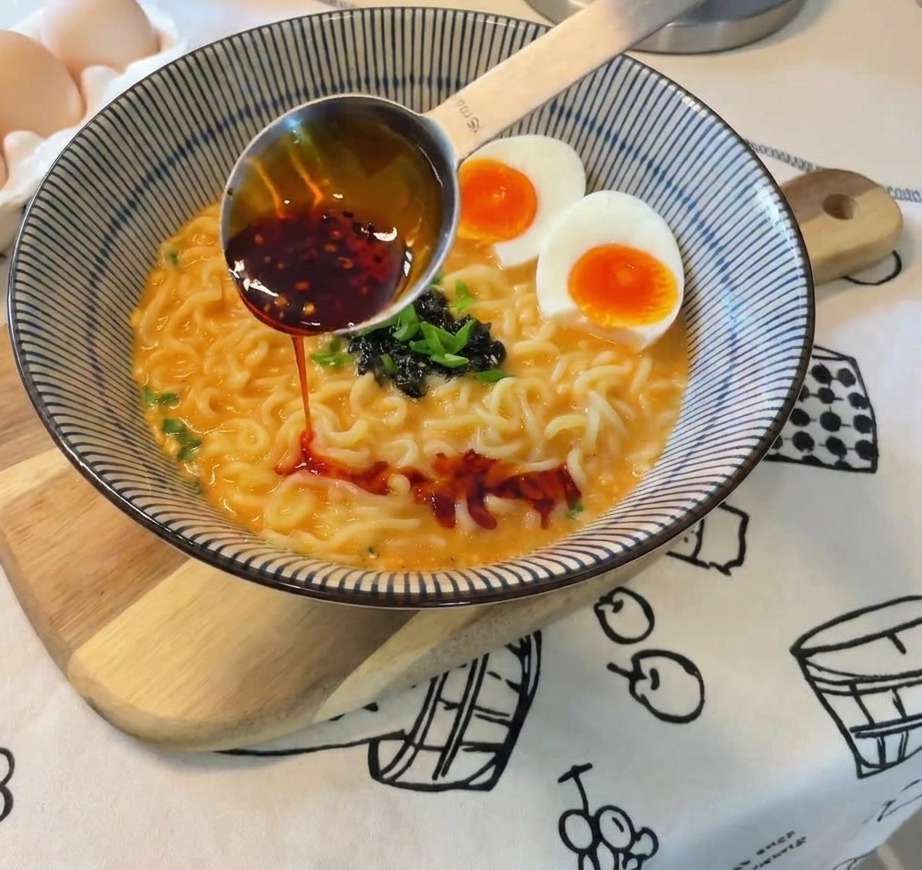 Aceite de chile rojo vibrante vertiéndose desde una cuchara de medición sobre el ramen terminado.