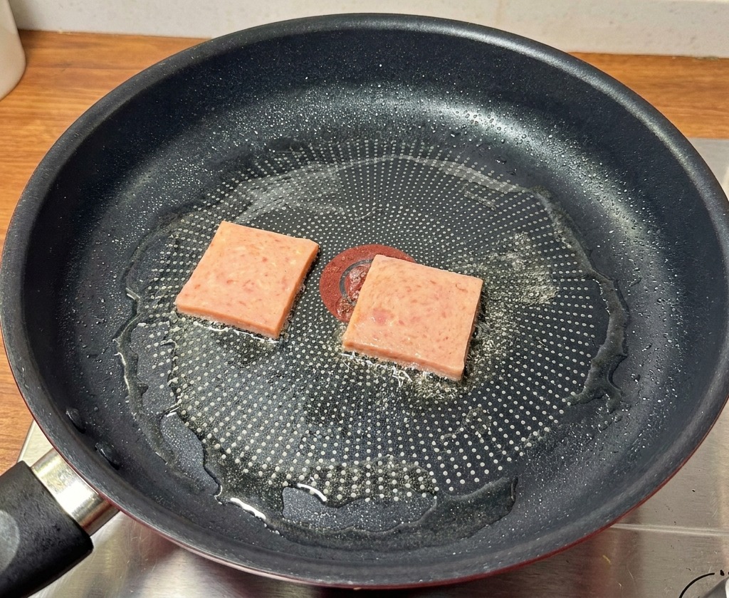Rebanadas de carne de almuerzo sellándose en una sartén negra hasta mostrar marcas doradas y crujientes.