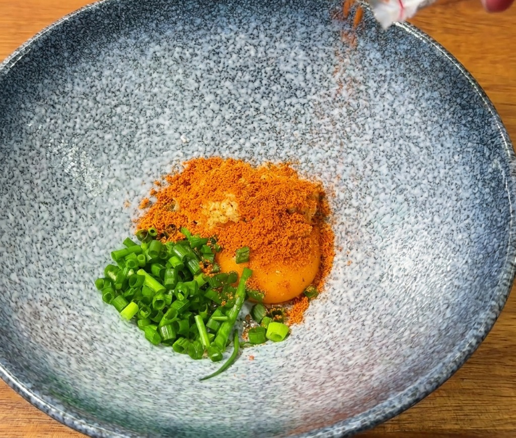 Sazonador de ramen picante vertiéndose en un bol azul que contiene una yema de huevo y cebolletas frescas.