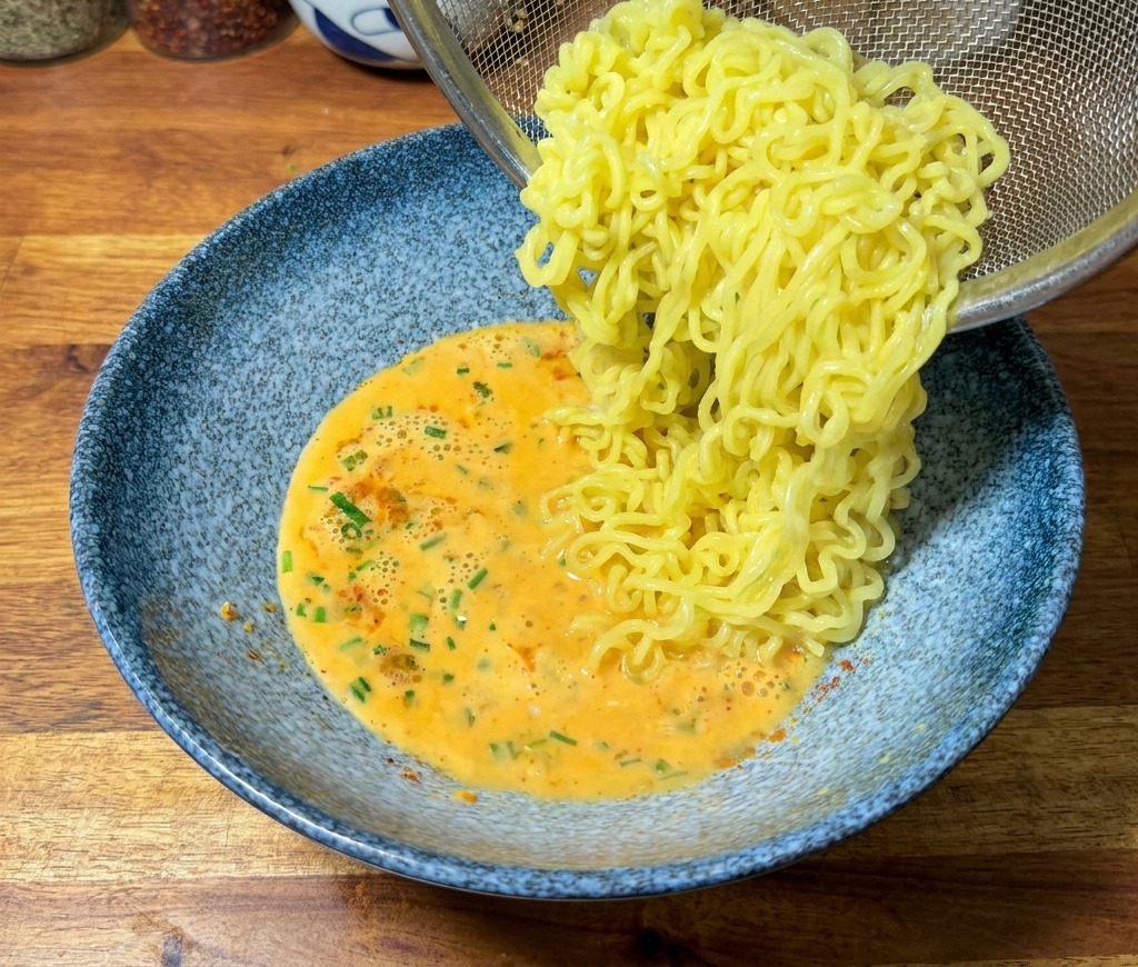 Fideos ramen amarillos vertiéndose desde un colador en un bol azul con caldo de color naranja cremoso.