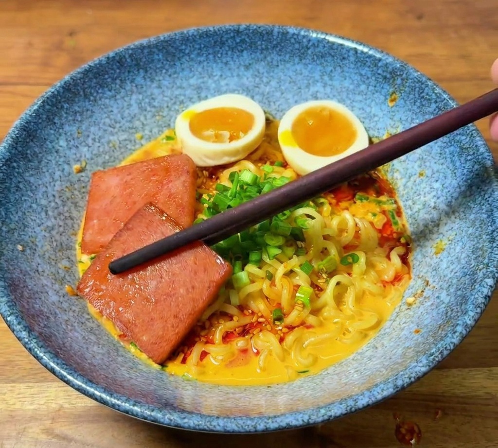 Unos palillos colocando una rebanada de carne de almuerzo dorada en el borde de un bol de ramen decorado.