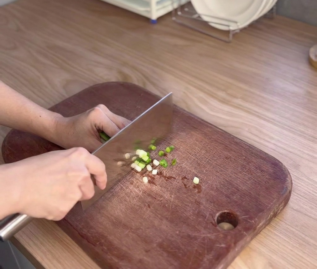 Una persona usa un cuchillo de chef grande para cortar finamente cebolletas frescas sobre una tabla de cortar de madera oscura.