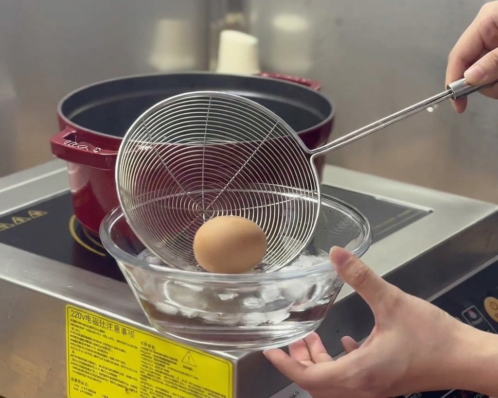 Un huevo cocido es trasladado con un colador desde una olla humeante a un cuenco de cristal con agua y cubitos de hielo.