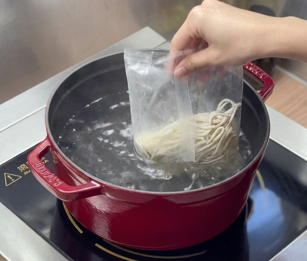 Fideos de ramen frescos siendo vertidos desde una bolsa de plástico transparente en una olla roja con agua hirviendo.