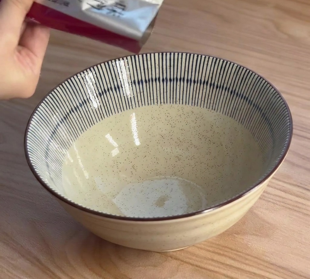 Una mano exprime una pasta espesa de condimento para ramen de miso desde un paquete plateado hacia un tazón de cerámica a rayas azules y blancas.