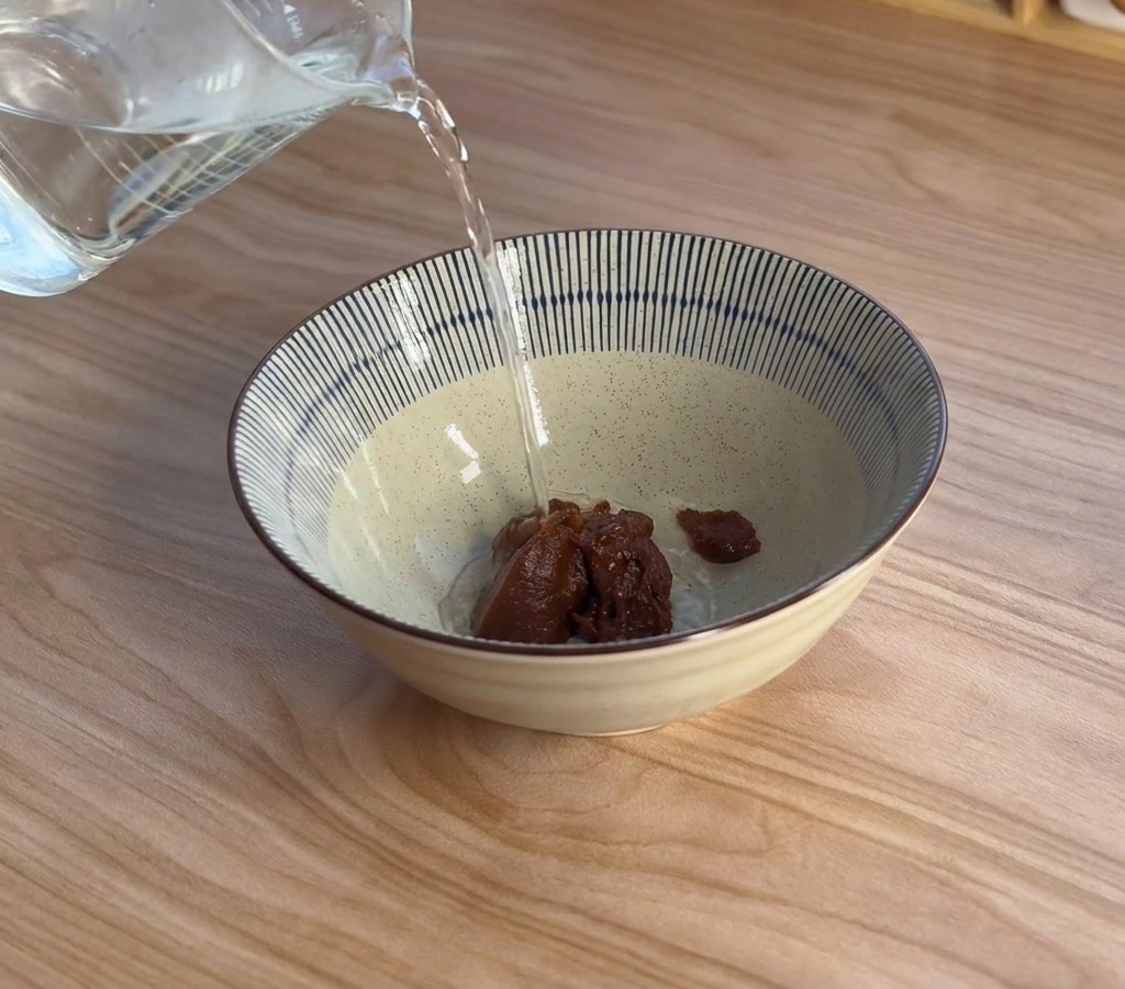 Vapor ascendiendo mientras se vierte agua hirviendo desde una jarra medidora sobre un cuenco de cerámica que contiene pasta de miso oscura.