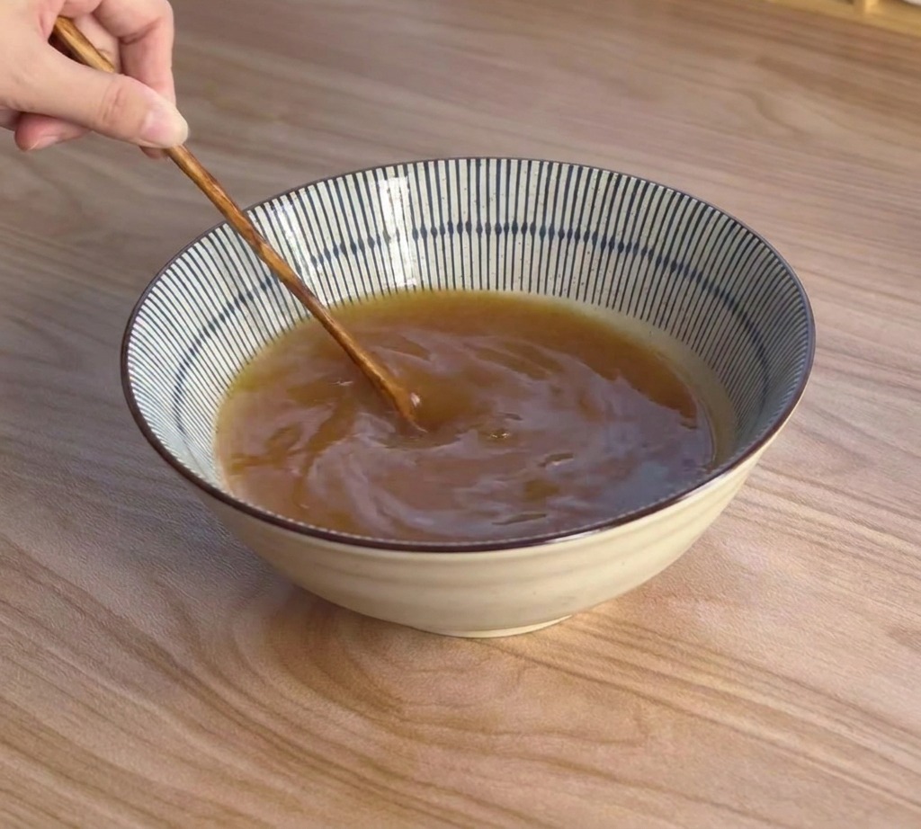 Una mano usando una cuchara de madera de mango largo para remover un caldo rico y amarronado en un tazón de cerámica a rayas.