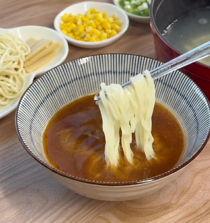 Fideos cocidos de ramen añadiéndose y revolviéndose dentro de un tazón con caldo de miso marrón usando palillos.