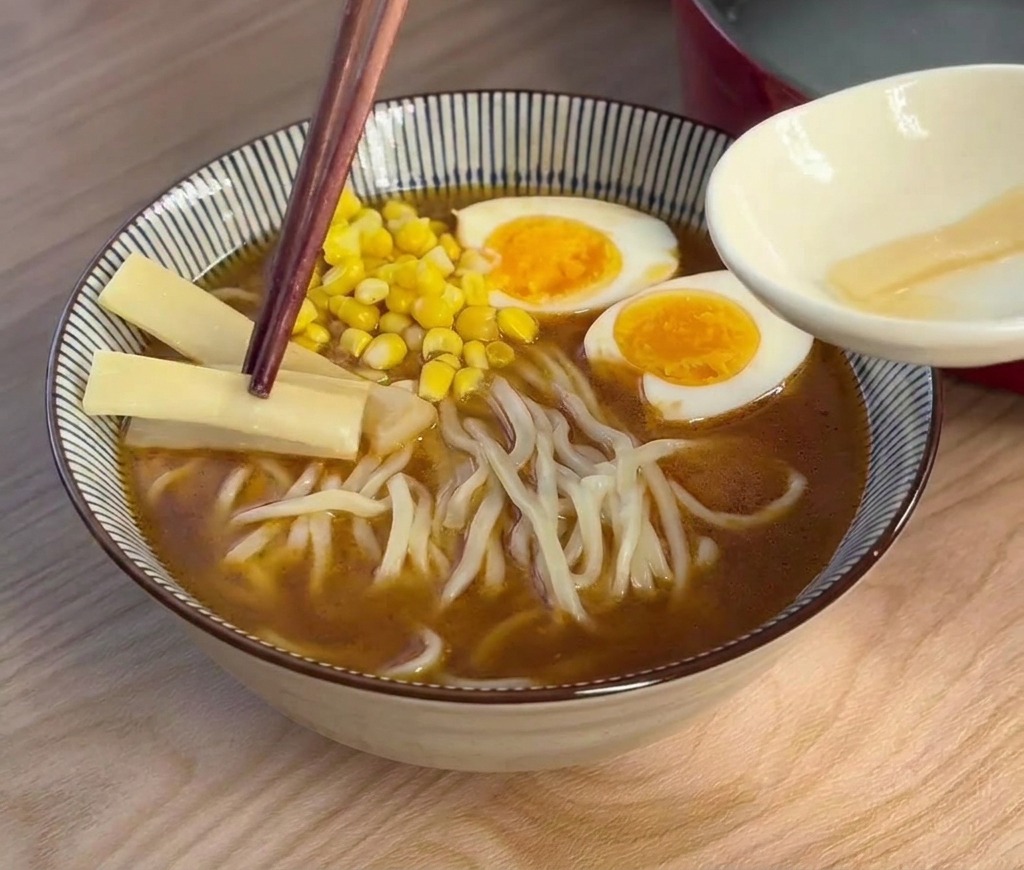 Palillos de madera oscura colocando tiras finas y claras de brotes de bambú en un ramen coronado con huevo y maíz.