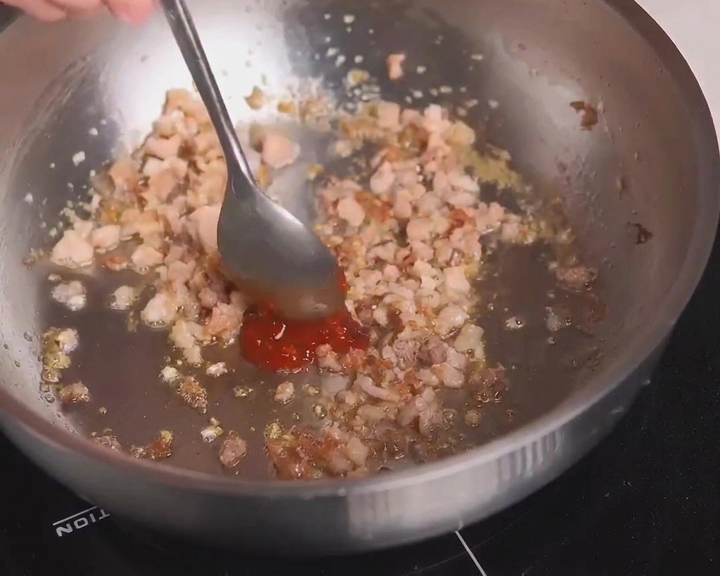 Una cuchara metálica añadiendo pasta de frijoles picante roja a la carne dorada.