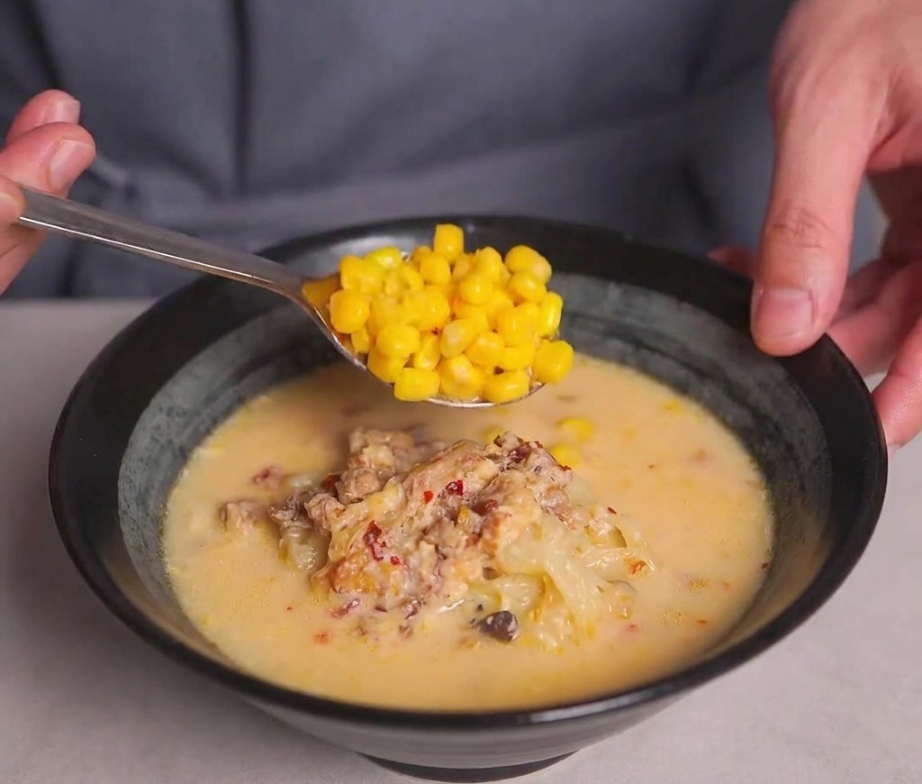 Una cuchara depositando maíz dulce dorado sobre el ramen cremoso.
