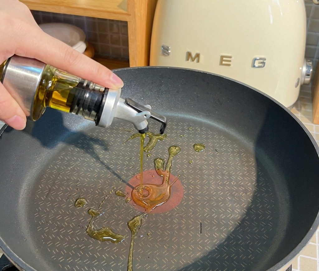 Aceite de cocina vertiéndose desde un dispensador de cristal en una sartén antiadherente negra.