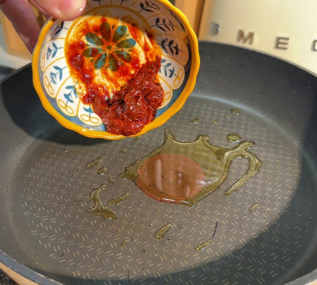 Una mano vertiendo pasta de judías roja picante desde un pequeño bol hacia la sartén con aceite caliente.