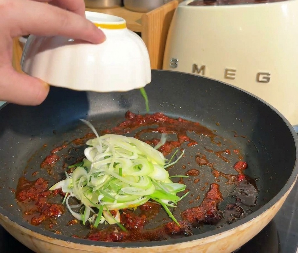 Cebolletas cortadas siendo añadidas a una sartén que contiene una base de salsa roja picante.