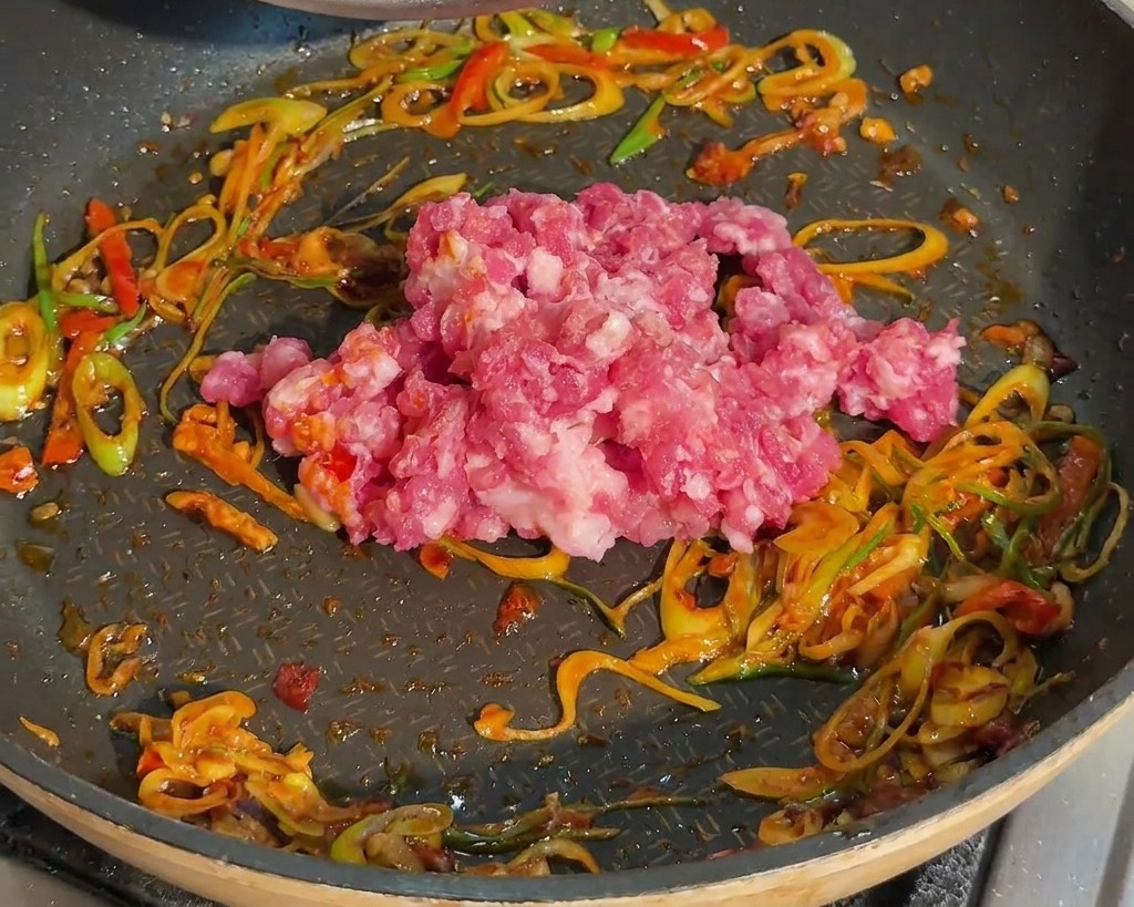 Carne de cerdo picada cruda colocándose en una sartén que ya tiene cebolletas y pasta de judías roja.
