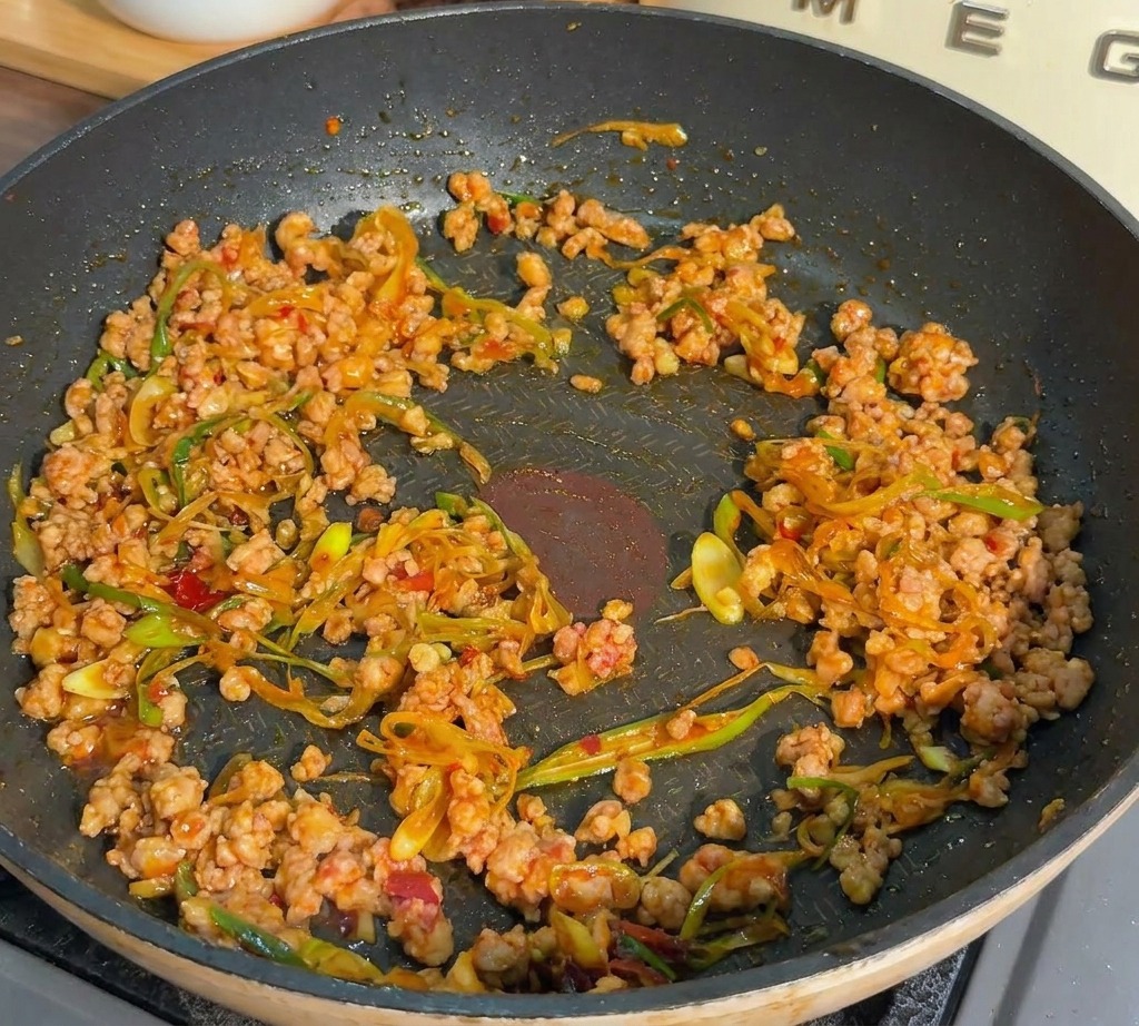Carne de cerdo picada y cebolletas salteándose en una sartén oscura hasta que la carne se ve dorada y cocinada.