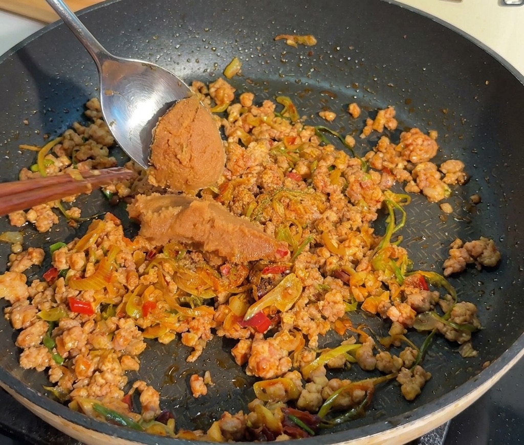 Una cuchara de metal añadiendo una porción de pasta de miso marrón oscuro a la sartén con carne cocinada.
