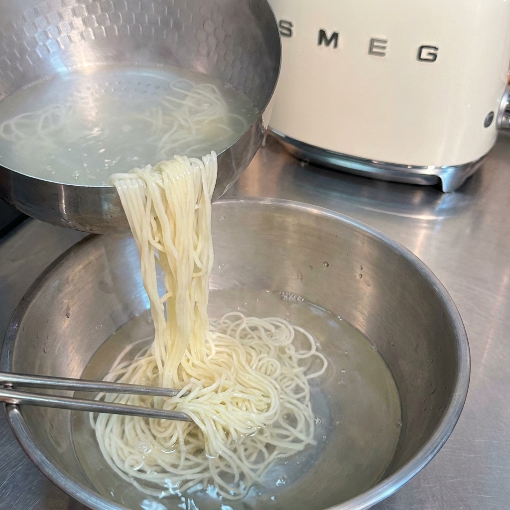 Fideos ramen cocinados siendo enjuagados con palillos en un bol de acero inoxidable con agua fría.