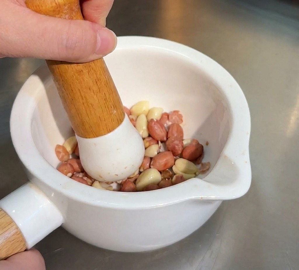 Una persona usando un mazo de madera para triturar cacahuetes tostados y semillas de sésamo en un mortero.